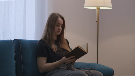 young-charming-lady-is-sitting-alone-in-living-room-and-reading-book-at-evening-turning-off-floor-lamp-calm-home-entertainment-at-weekends